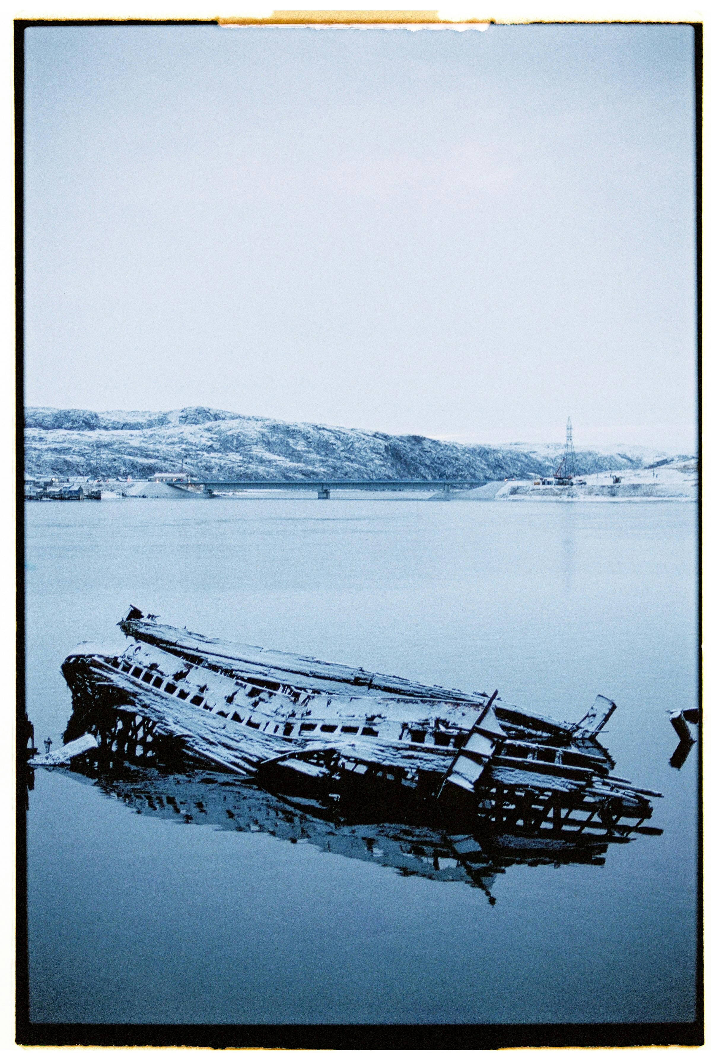 A serene winter scene of a shipwreck in the icy waters of Териберка, Russia, surrounded by snow-covered hills.