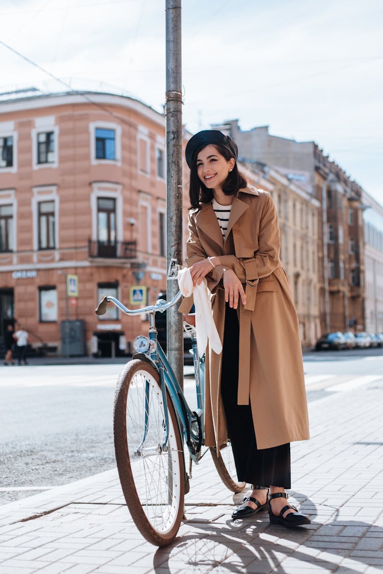 A Woman In Brown Coat Standing On The Street Near Her Bike