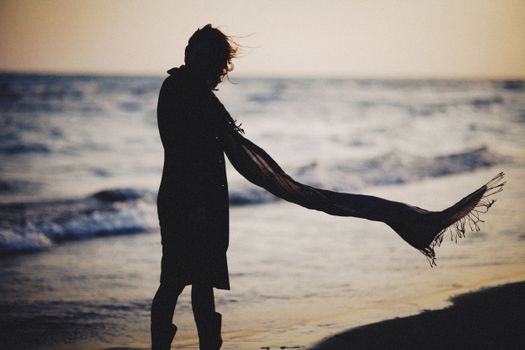 A silhouetted figure with a scarf by the sea during sunset, capturing a serene beach moment.