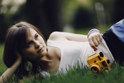 Young woman in a white tank top lying on grass, holding a vintage camera with a thoughtful expression.