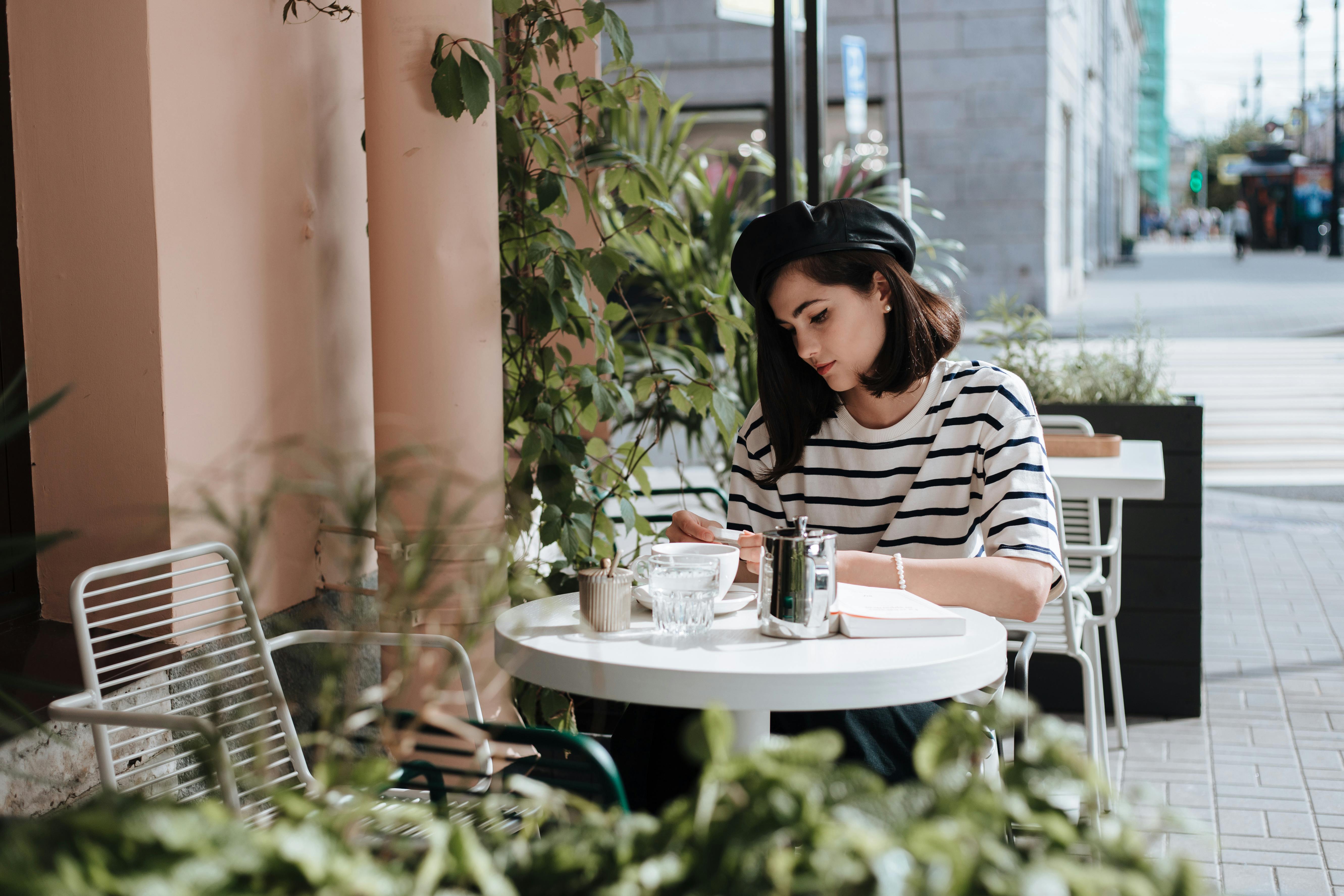 Free Elegant woman in beret reading at a charming outdoor café with plants. Stock Photo