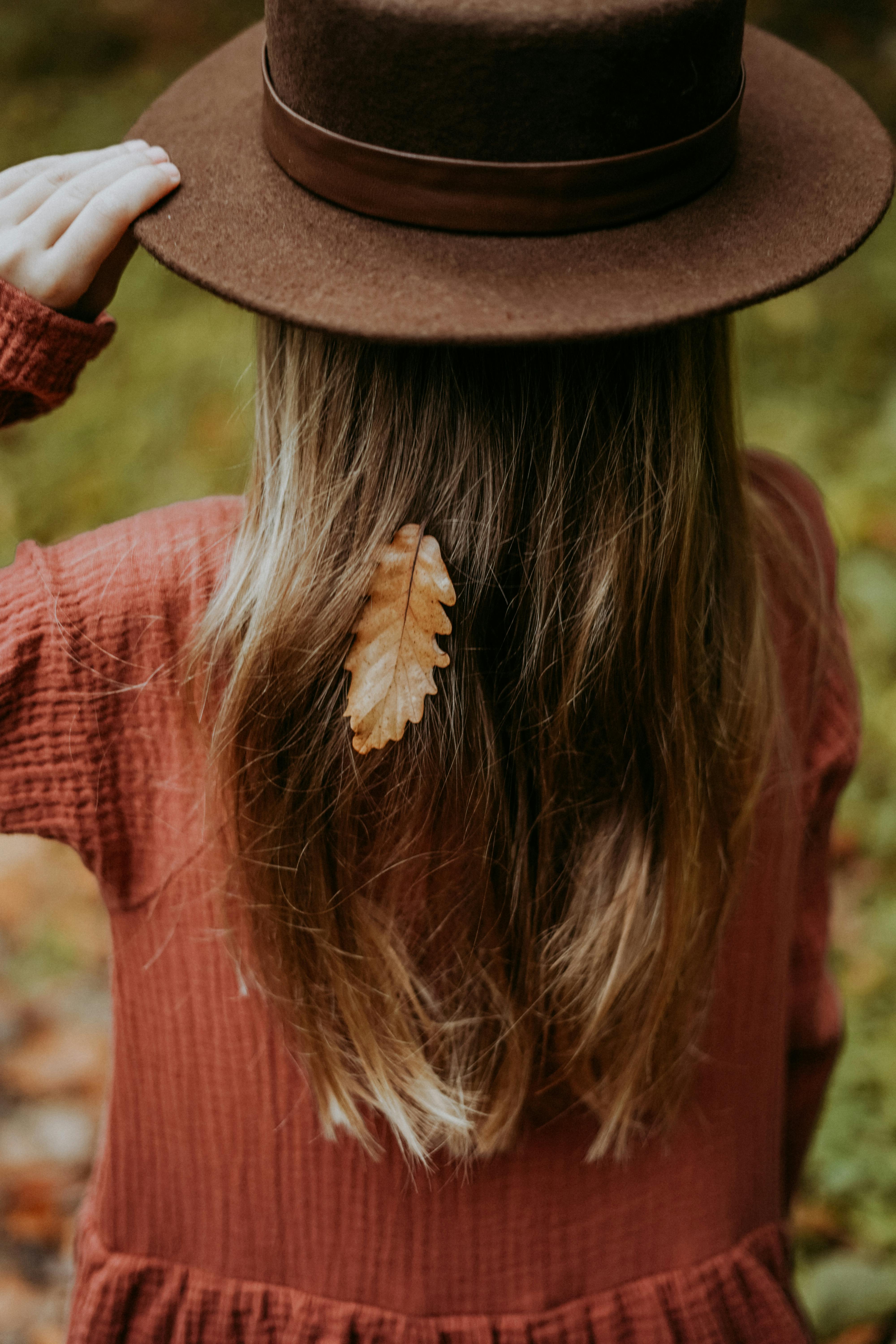 Back view of a girl with long brown hair, wearing a hat, holding a leaf on her autumn dress.