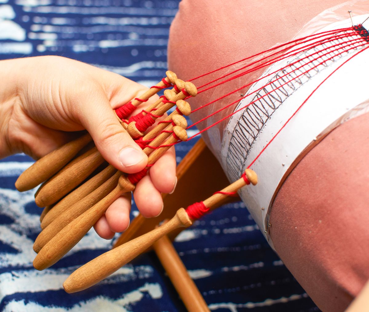 Detailed view of hands crafting bobbin lace with wooden tools and red threads.
