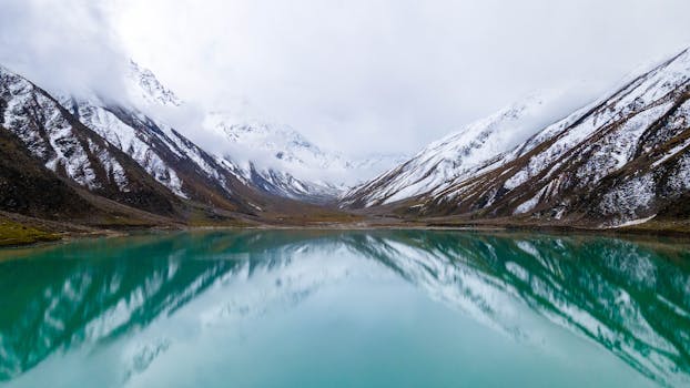 Breathtaking view of snow-capped mountains mirrored in a clear, turquoise lake under soft, cloudy skies.