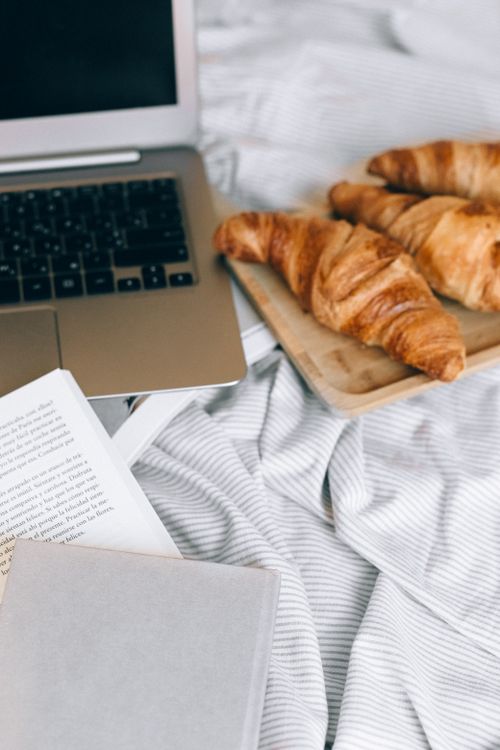 Free Cozy breakfast in bed with croissants, open book, and a laptop for a peaceful morning. Stock Photo
