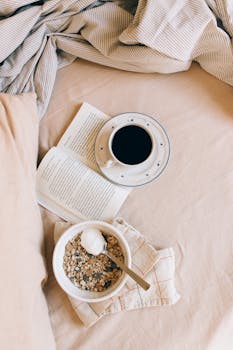 Top view of a comforting breakfast with black coffee, granola, and an open book on a cozy fabric backdrop.