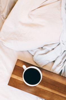 A warm cup of black coffee resting on a wooden tray beside a comfortable bed.