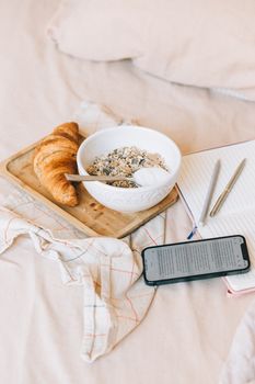 A tray of croissant and yogurt with granola on a bed, accompanied by a notebook and smartphone.