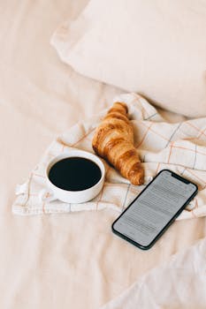 Flat lay of a cozy breakfast scene featuring a croissant, coffee, and smartphone on a blanket.