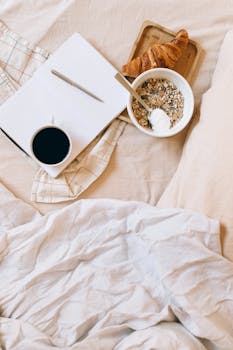 A warm and inviting breakfast setup with coffee, croissant, and yogurt granola bowl on a comfy bed.