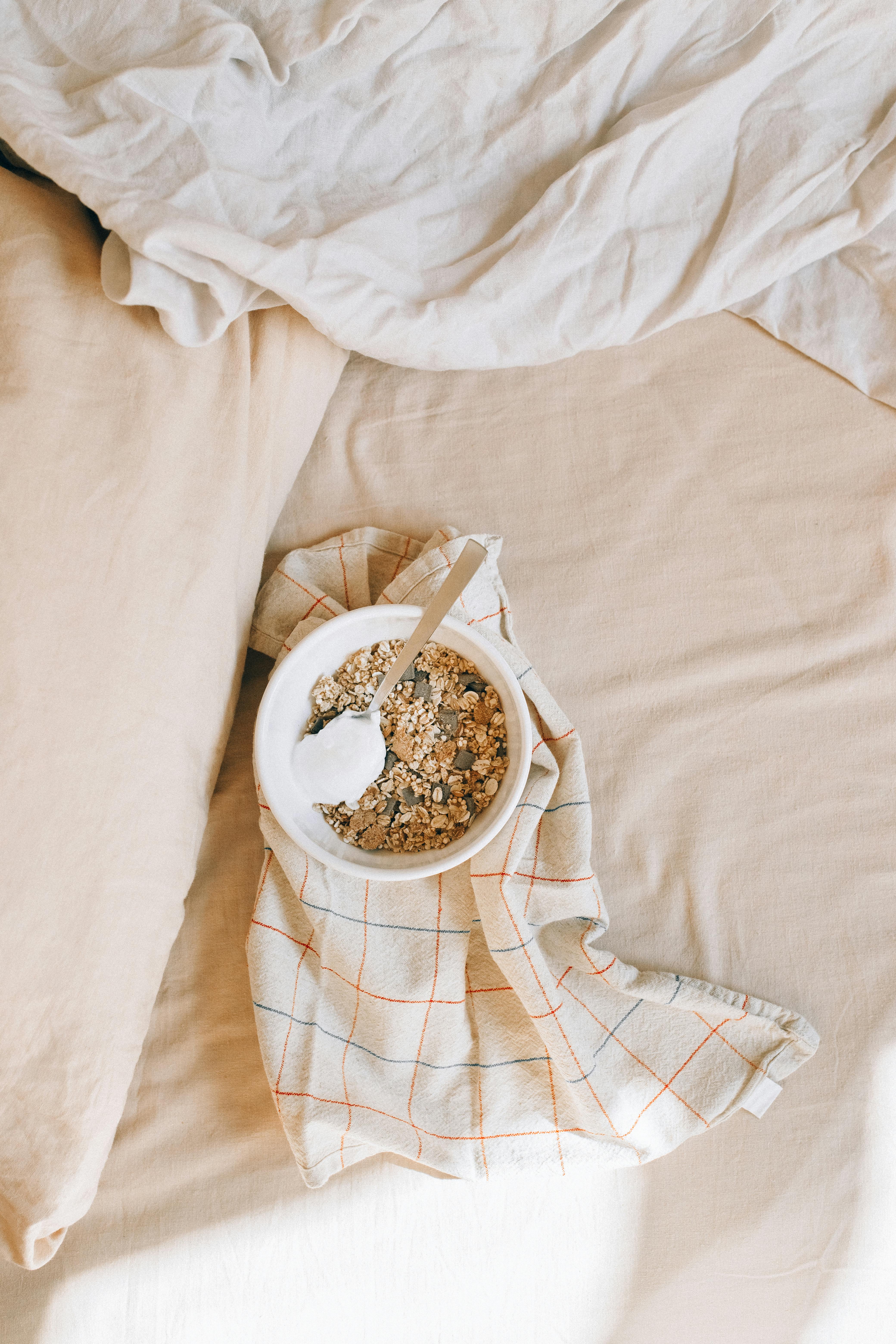 A bowl of granola with yogurt on a plaid kitchen towel in a cozy bed setting, viewed from above.