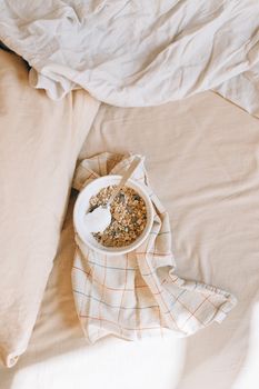 A bowl of granola with yogurt on a plaid kitchen towel in a cozy bed setting, viewed from above.