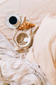 Warm morning breakfast setup with coffee, croissant, and cereals on a bed.
