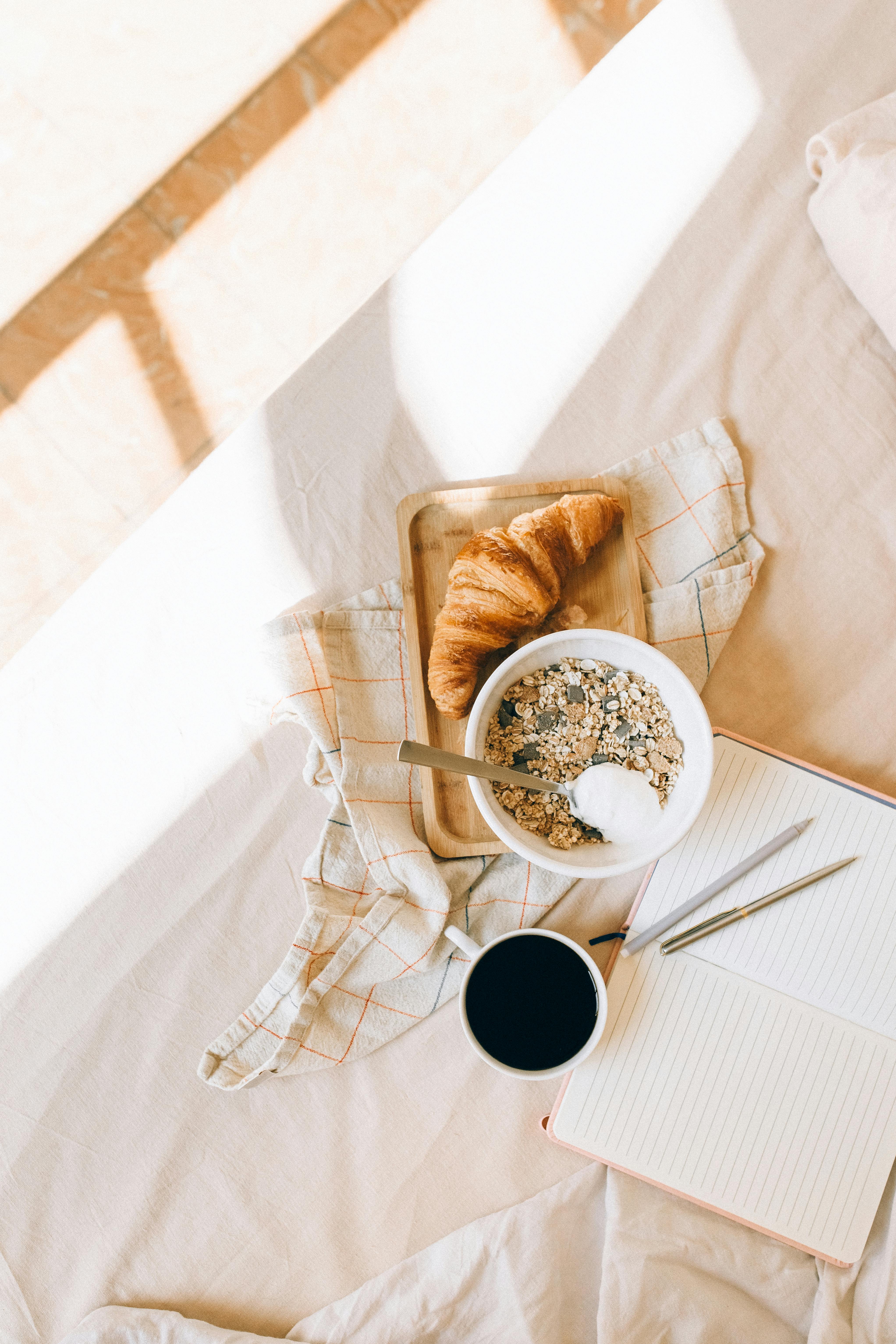 Flat lay of cozy breakfast with croissant, coffee, and notebook on bed.