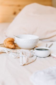 Stylish breakfast setup on bed with a croissant, coffee, and a bowl, evoking a warm and cozy morning.