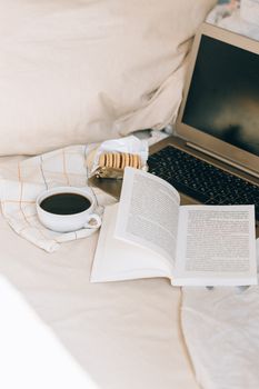 A cozy bed setting featuring a laptop, open book, coffee, and cookies for a relaxing reading session.