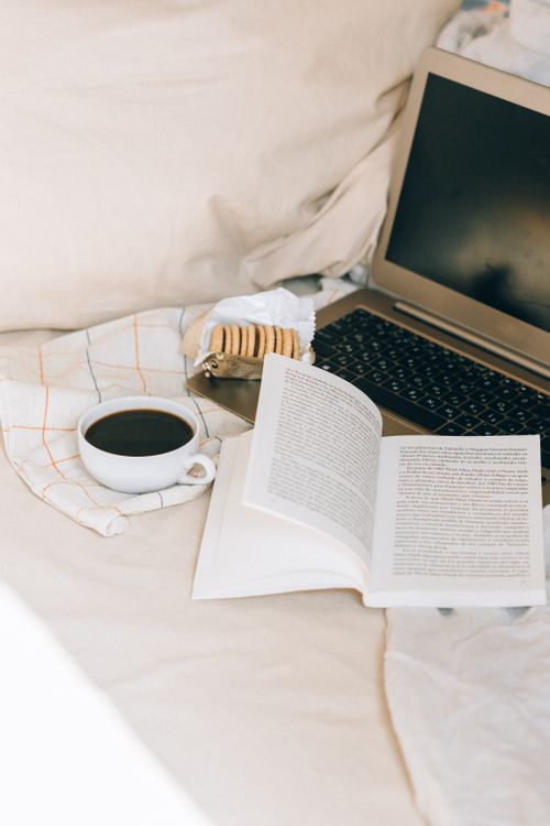 Free A cozy bed setting featuring a laptop, open book, coffee, and cookies for a relaxing reading session. Stock Photo