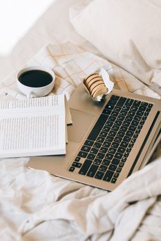 Comfortable morning setup with coffee cup, open book, and laptop on a bed.