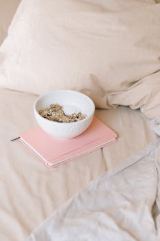 Bowl of cereal on a pink notebook in a cozy bed setting, perfect for a relaxing morning.