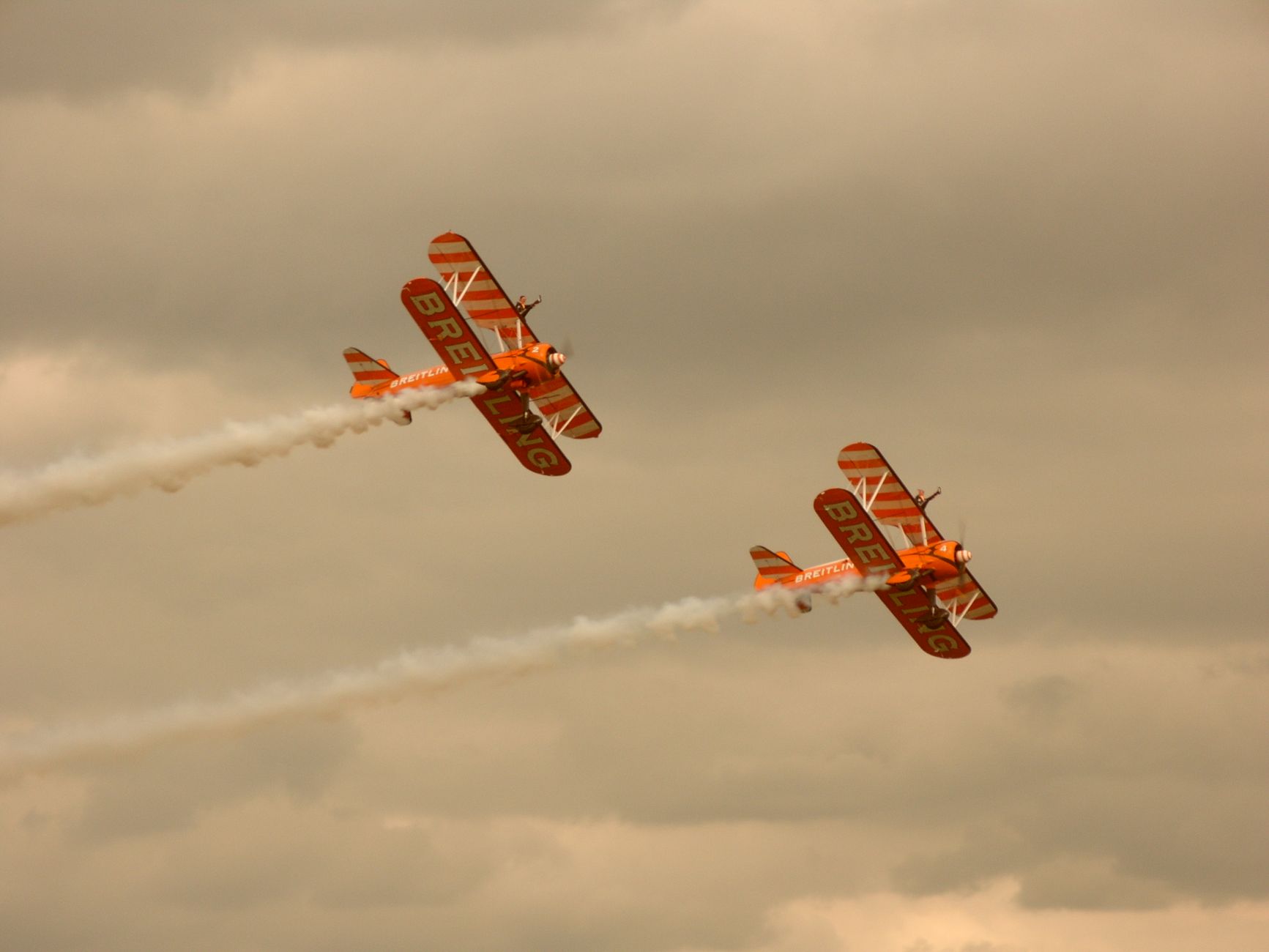 Two classic biplanes perform aerial stunts under cloudy skies, leaving smoke trails.