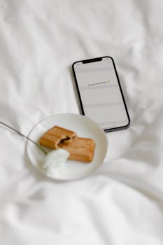 A cozy morning setup with a smartphone, biscuits, and a flower on a white blanket.