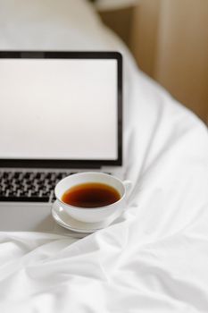 A laptop with a blank screen and a cup of coffee on a cozy white bed setting.