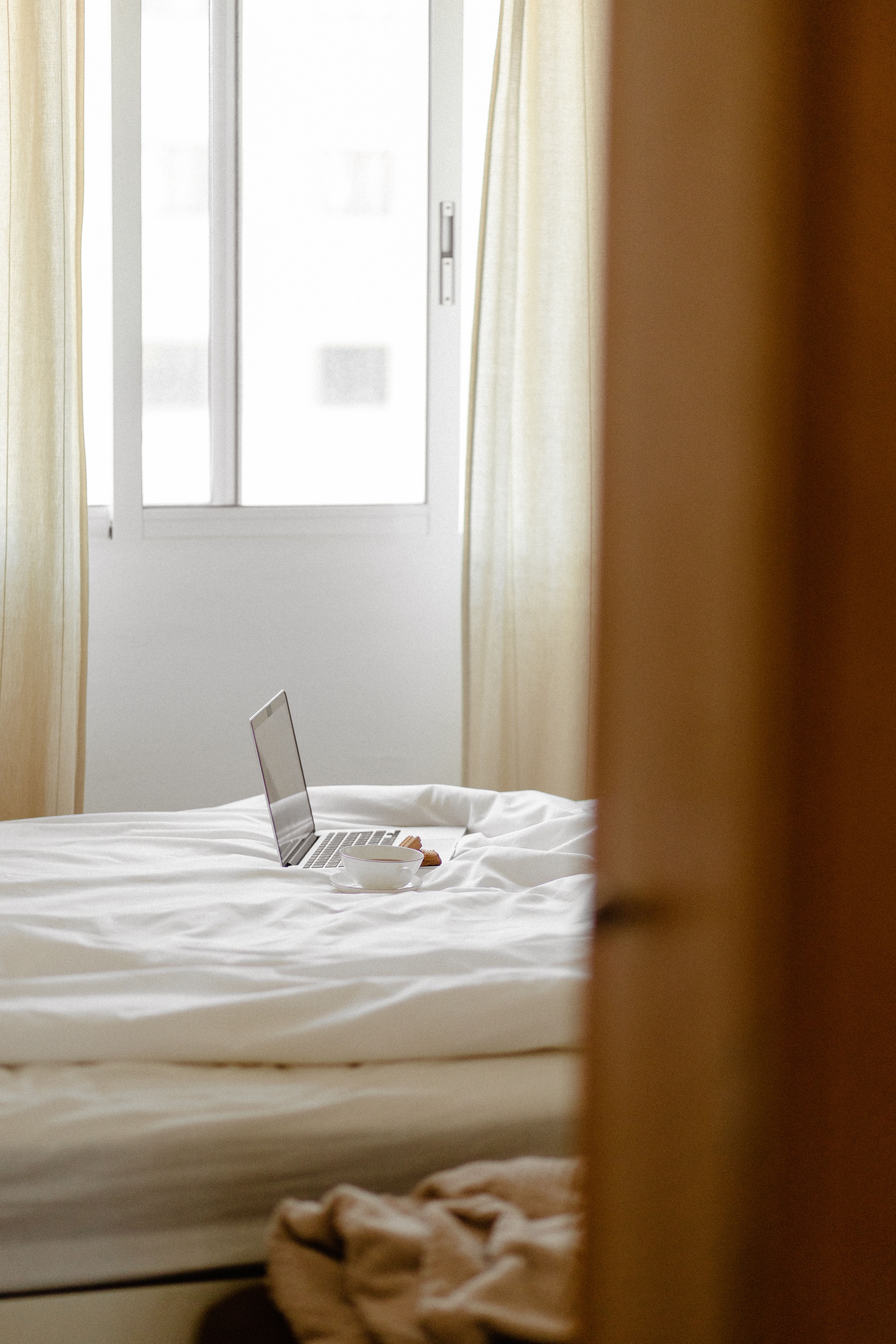 A cozy bedroom scene with a laptop and coffee cup on a bed next to a window.
