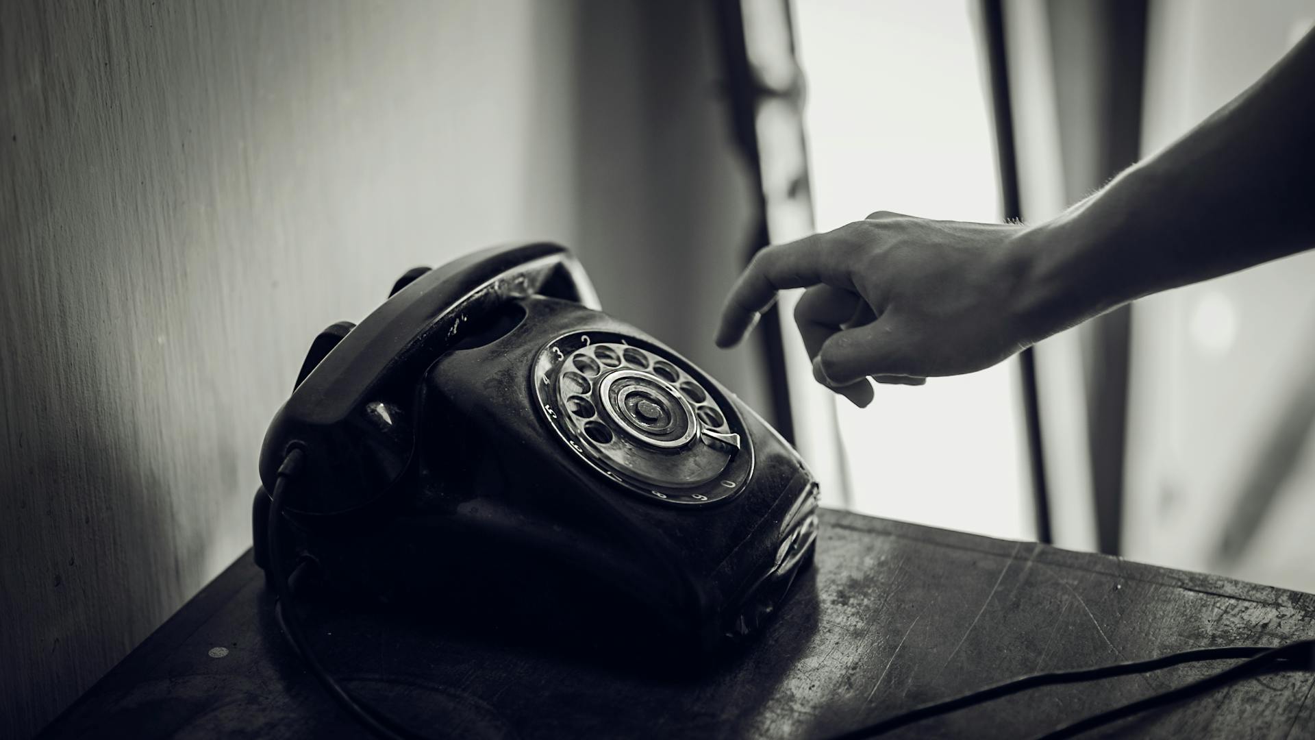 A monochrome photo of a hand reaching towards a vintage black rotary telephone in an indoor setting.
