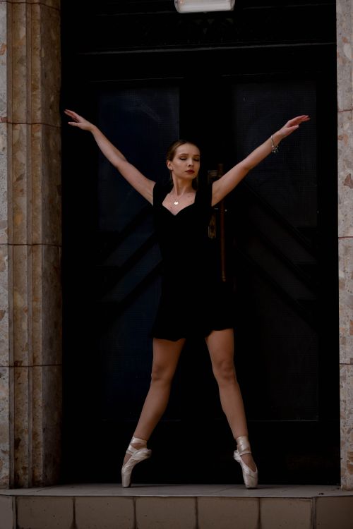 Free Ballerina in black dress gracefully poses on pointe shoes outside a dark doorway. Stock Photo