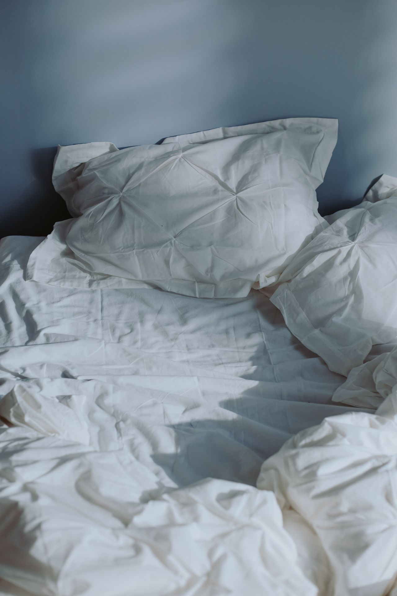 Unmade white bed with pillows against a blue wall in morning light
