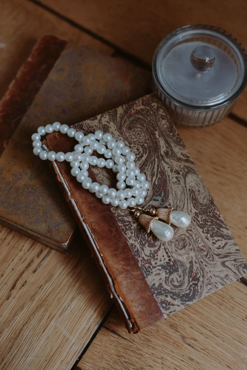 Free Elegant pearl necklace resting on a vintage book with a glass jar, creating a classic still life arrangement. Stock Photo