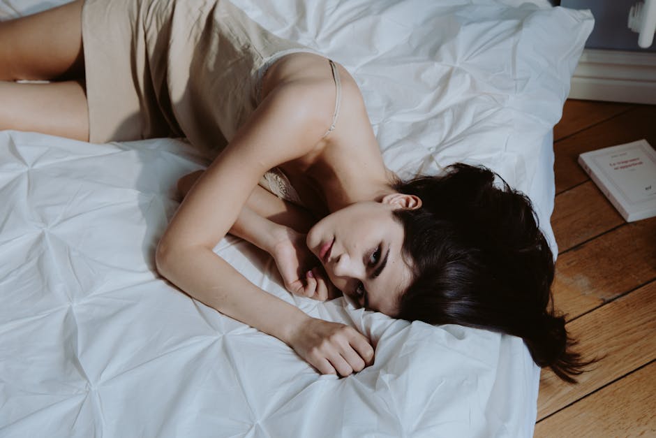 Tânără cu păr lung întinsă pe pat alb, cufundată în lumină moale care intră prin fereastra dimineții