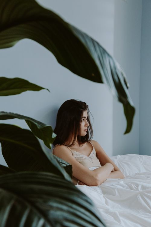 Free A contemplative woman sits cross-armed on a bed, framed by lush green leaves in a serene bedroom. Stock Photo
