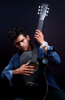 Young man posing with an acoustic guitar against a dark background, showcasing musical talent.