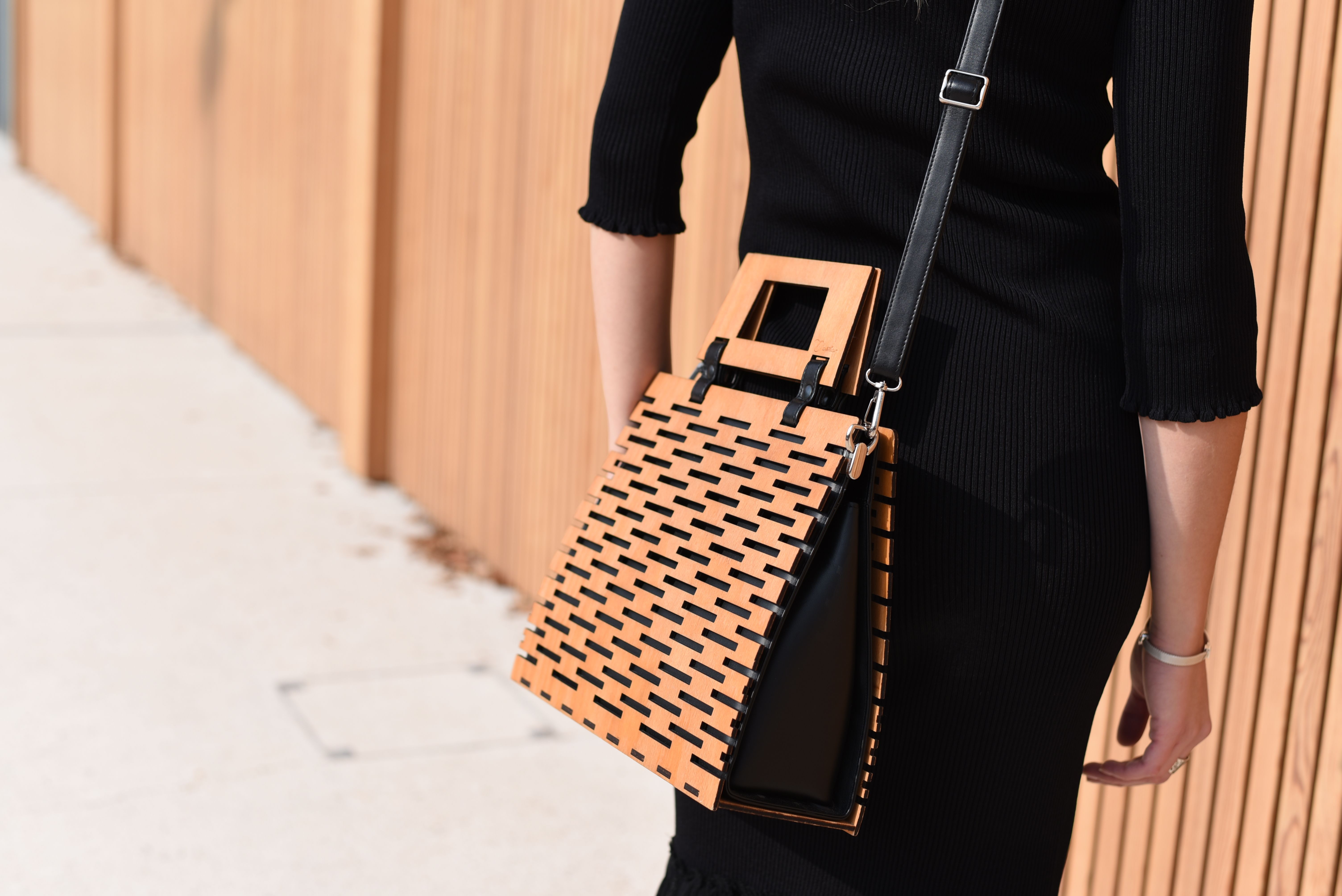 Fashionable woman in black dress showcasing a unique woven handbag against an urban backdrop.