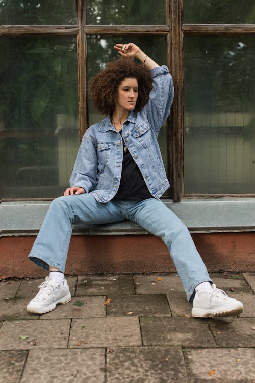Free A Woman in Denim Jacket and Pants Sitting on the Street Near the Glass Window with Wooden Frame Stock Photo