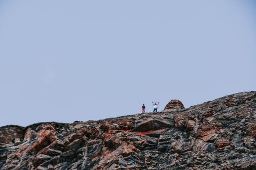 Free Two people stand triumphantly atop a rocky mountain peak under a clear blue sky. Stock Photo