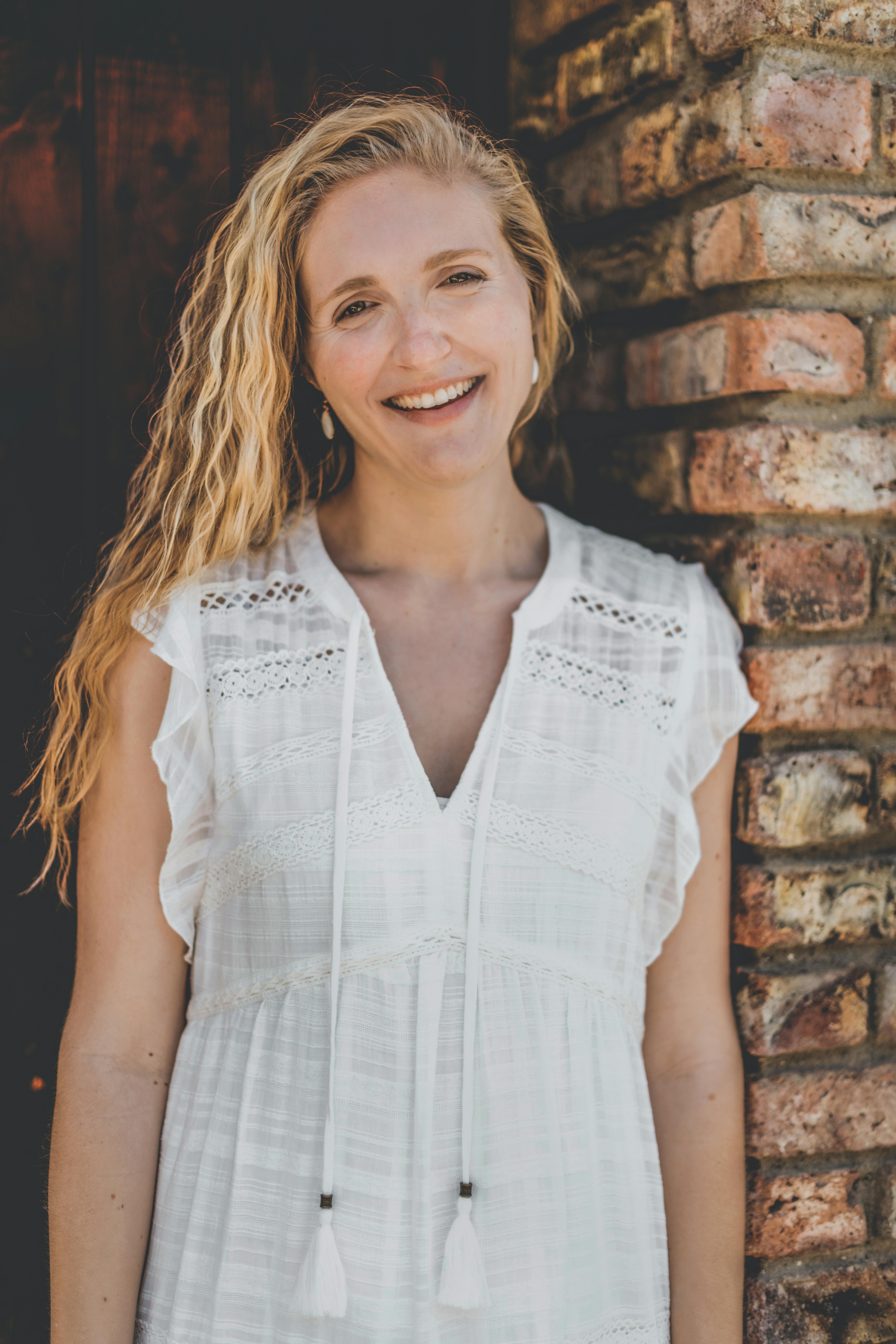 Casual portrait of a smiling woman in a white dress leaning against a brick wall.