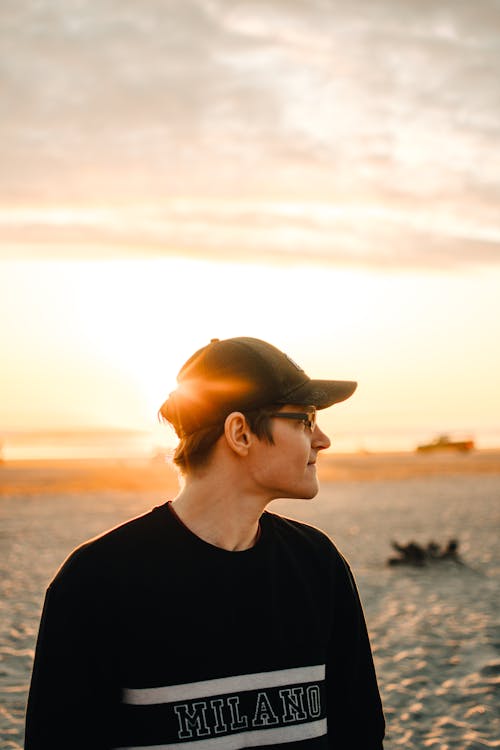 Free Man in Black Sweater and Black Cap Standing on Beach During Sunset Stock Photo