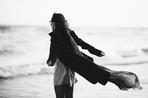 Free A woman in a coat stands by the ocean, her scarf blowing in the wind, captured in grayscale photography. Stock Photo