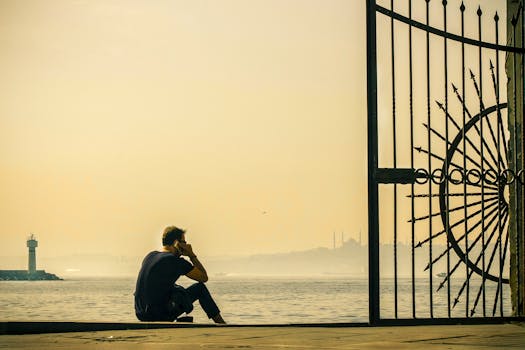 A contemplative man sits by the ocean at sunrise near an ornate gate, with a distant lighthouse in view.
