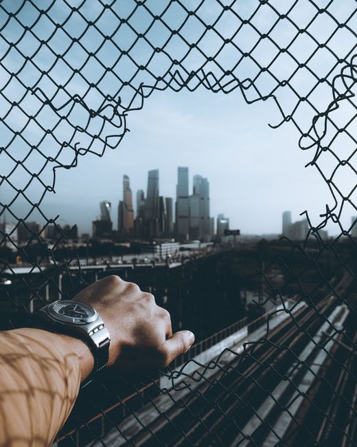 A hand with a watch reaches through a chain-link fence, overlooking Moscow's cityscape.