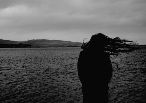 Free A moody black and white silhouette of a woman with flowing hair standing by the sea at twilight. Stock Photo