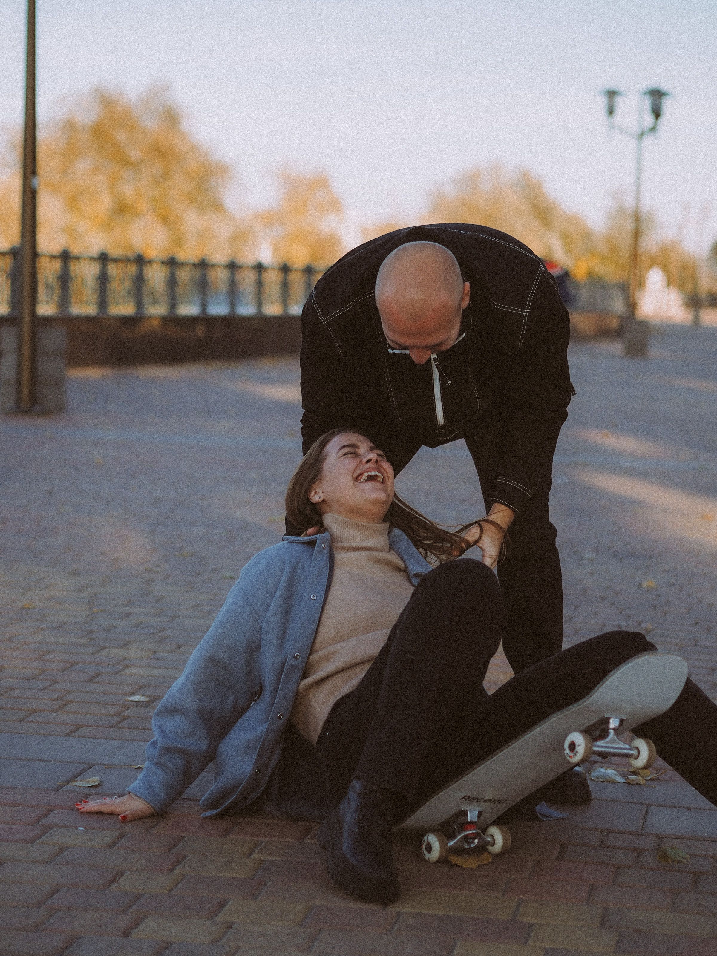 A couple shares a joyful moment on the pavement with a skateboard in an outdoor park setting.