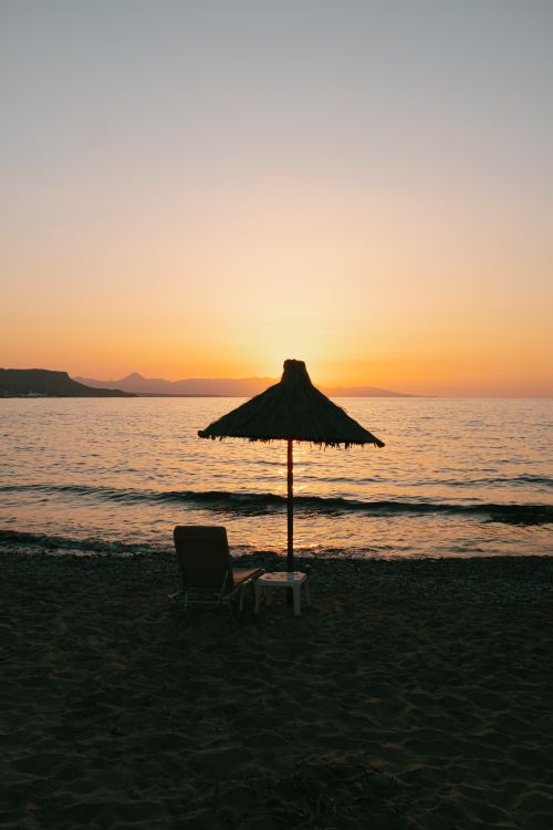 Free Peaceful beach scene at sunset in Greece with a silhouette of a straw hut and chair. Stock Photo