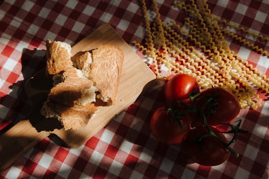 Artistic arrangement of bread, tomatoes, and pasta on a classic red-checkered tablecloth with warm sunlight.