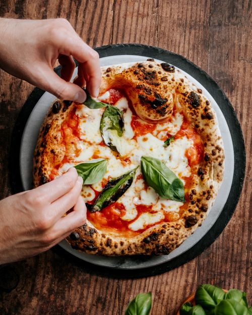 Free Close-up of hands adding basil to a freshly baked Neapolitan pizza, highlighting its cheesy and delicious appeal. Stock Photo