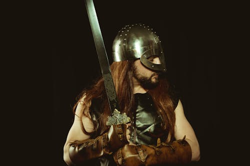 Long Haired Man in Metal Helmet and Leather Gloves Holding Silver Sword