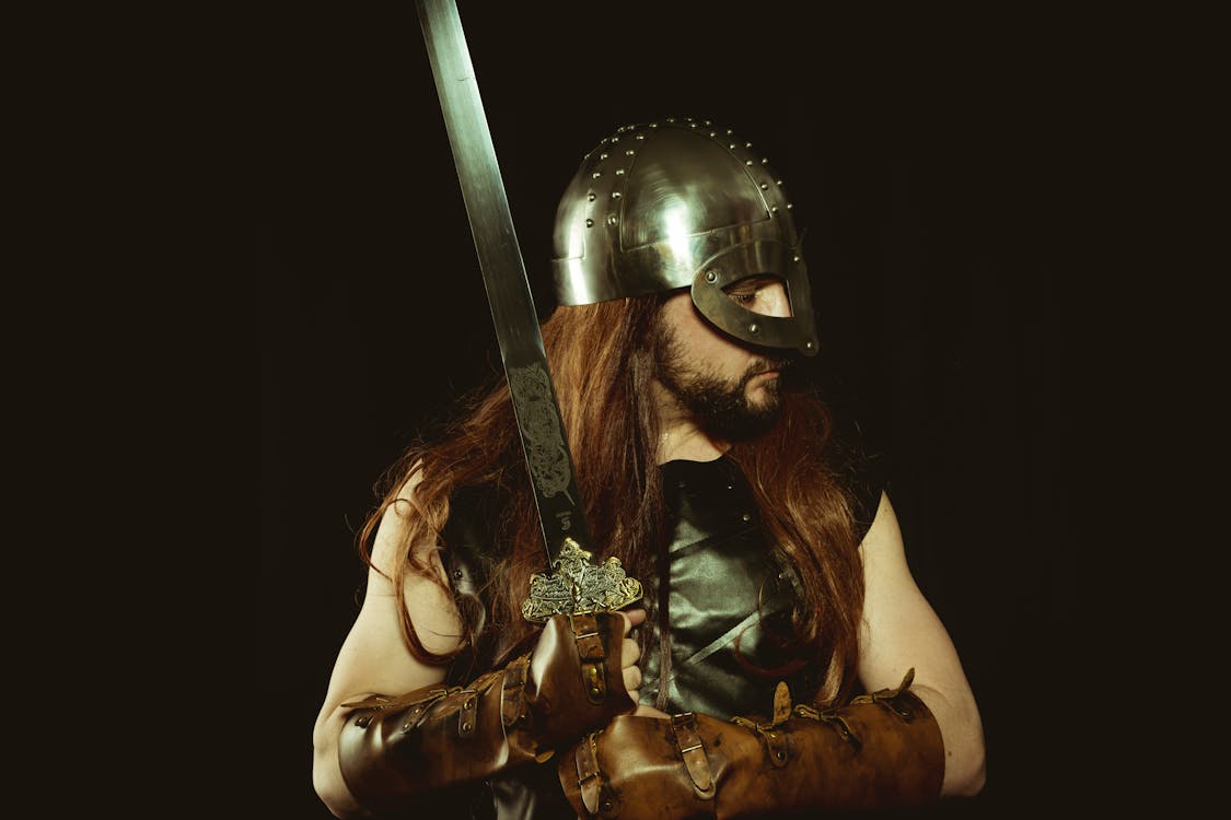 Long Haired Man in Metal Helmet and Leather Gloves Holding Silver Sword
