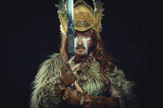 A dramatic portrait of a Viking warrior in costume holding a sword, featuring a golden headdress and fur garment.